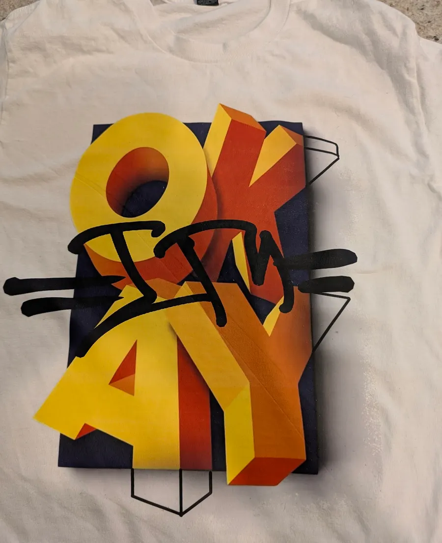 White T-shirt printed with a large yellow and orange OKAY graphic and black tag-style lettering.
